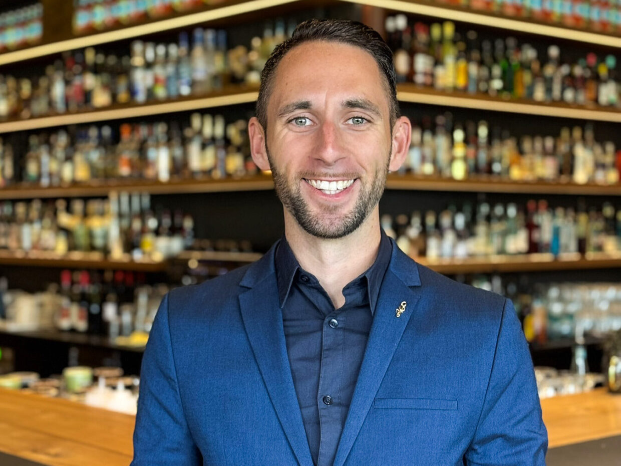 Man in blue jacket smiling in front of alcohol shelf