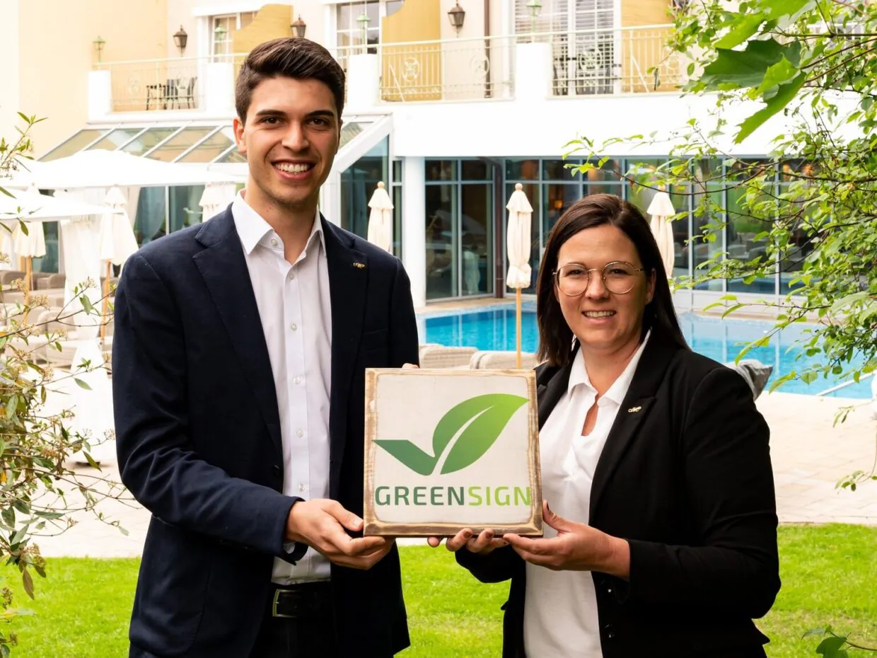 Zwei Personen halten Greensign-Logo vor Hotelpool.